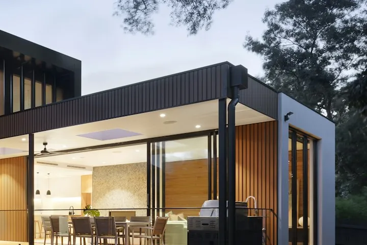 Covered outdoor dining terrace under a projecting roof, opening onto a lawn at dusk.