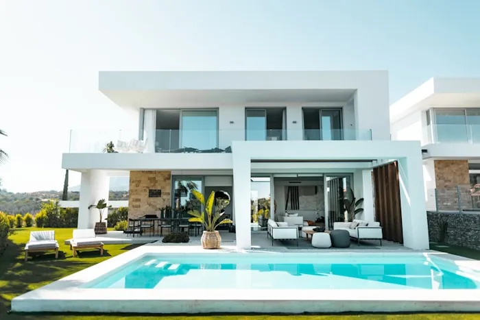 Covered outdoor lounge and dining area beside a pool, shaded by the projecting roof of a villa.