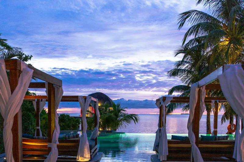 Draped timber shade structures beside water at sunset, framed by palms.