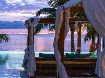 A close view of one timber cabana frame with white drapes tied back, palms and open water behind.