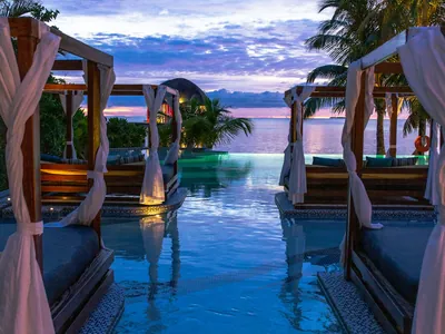 Two draped timber cabanas standing either side of a lit pool, looking out to open water at sunset.