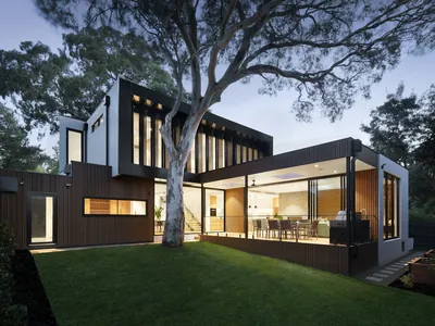 A level lawn running up to the lit glazed elevation of a dark-clad single-storey house at dusk, with a large tree alongside.