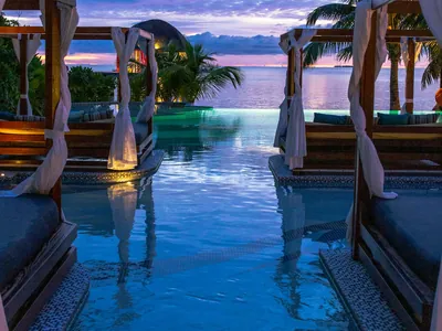 A lit pool edge meeting open water at dusk, with a draped cabana and palms alongside.