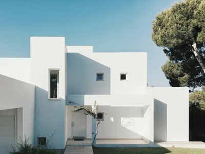 Stepped white rendered volumes with a deep shaded recess, seen against a clear sky with a pine behind.