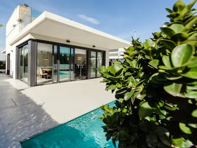 Wide sliding glazed doors opening from a single-storey house onto a paved terrace beside a pool.