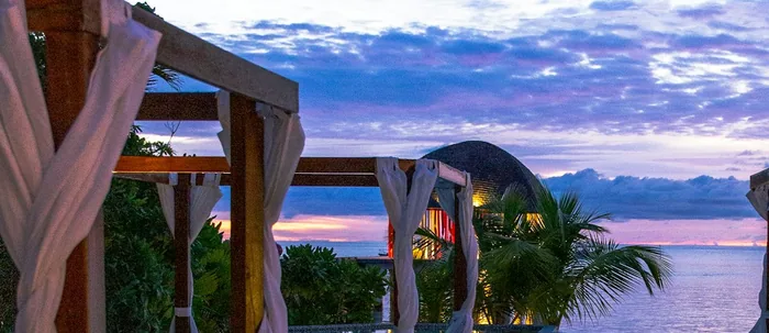 Timber post-and-beam shade structure with fabric drapes beside water at sunset.